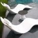 Two modern white wavy lounge chairs with cushions arranged around a small round dark gray side table on a dark pool deck next to a swimming pool.