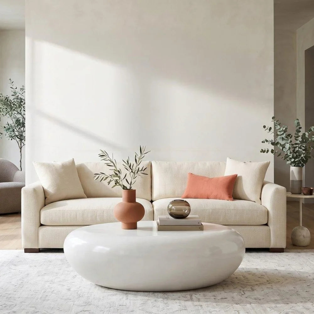 A cream-colored fabric sofa in a bright, modern living room setting, styled with a terracotta vase, a small metallic orb, and a coral accent pillow, set behind a glossy white oval coffee table.