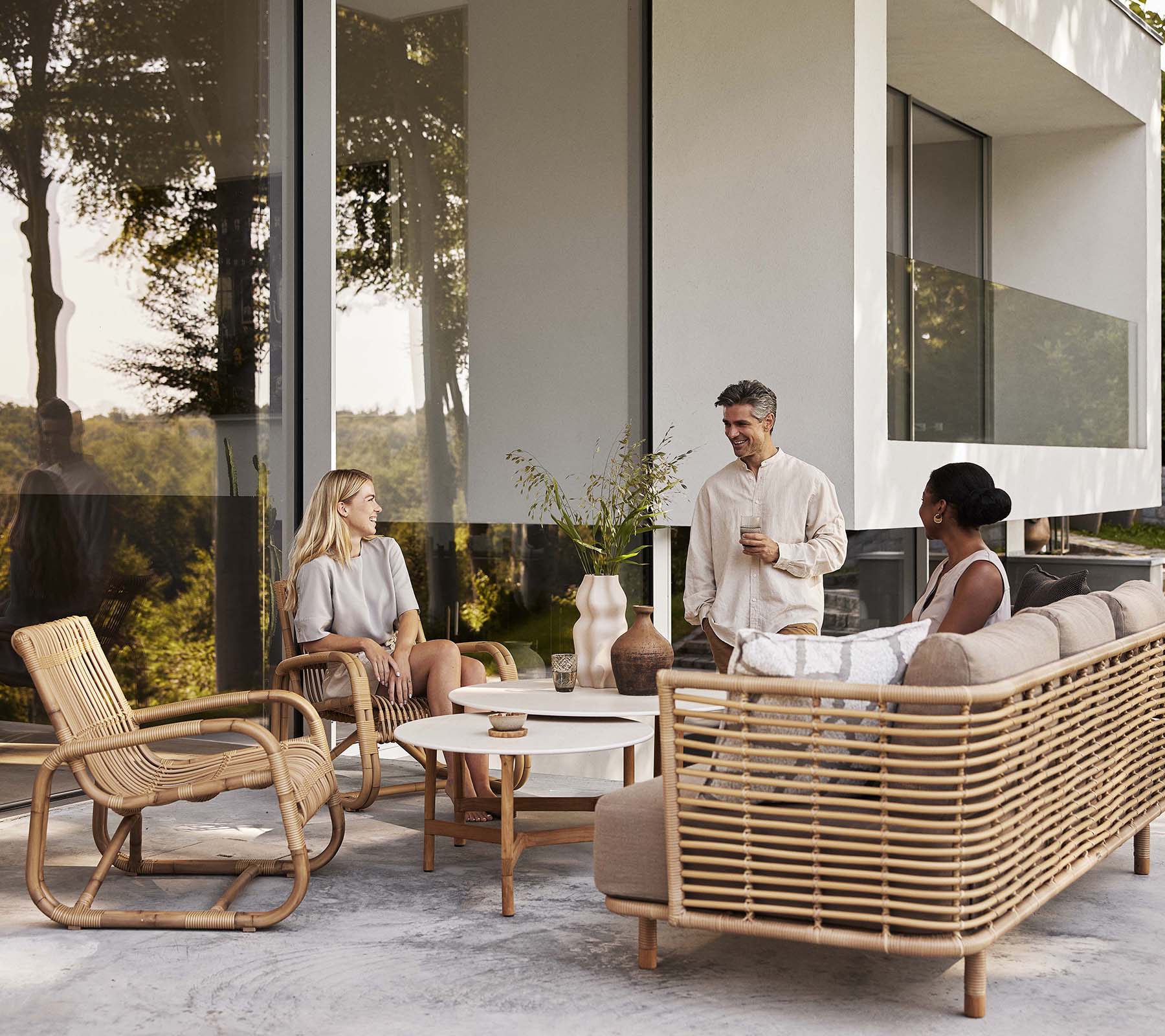 Boxhill's Curve Lounge Weave Outdoor Chair Natural with 2 round tables, 3-seater sofa, 2 women sitting down and a man standing beside