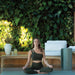 Woman meditating in a serene outdoor setting with greenery and a couch.
