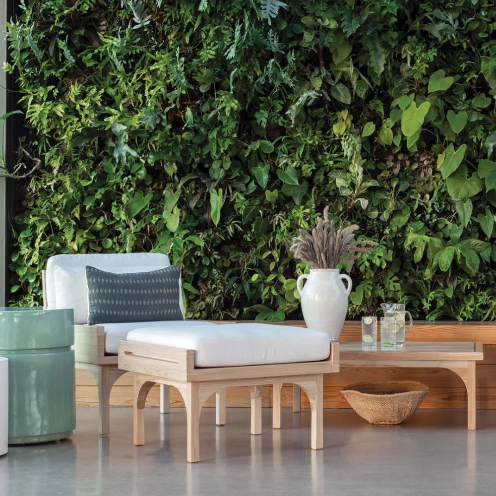 Outdoor seating area with a wooden bench, white cushions, and a vase against a green wall.