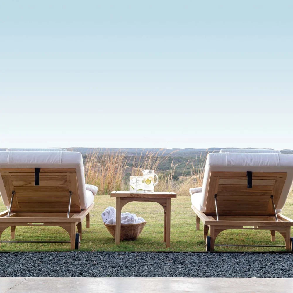 Two wooden outdoor lounge chairs with a small table in the middle on a grassy area with a scenic background.