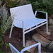 A white aluminum outdoor bench with slatted design placed in the garden surrounded with greenery