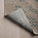 A close-up of a woven area outdoor rug with a curled corner, showing a textured beige and light blue pattern on the top side and a muted blue underside, placed on a neutral beige surface.