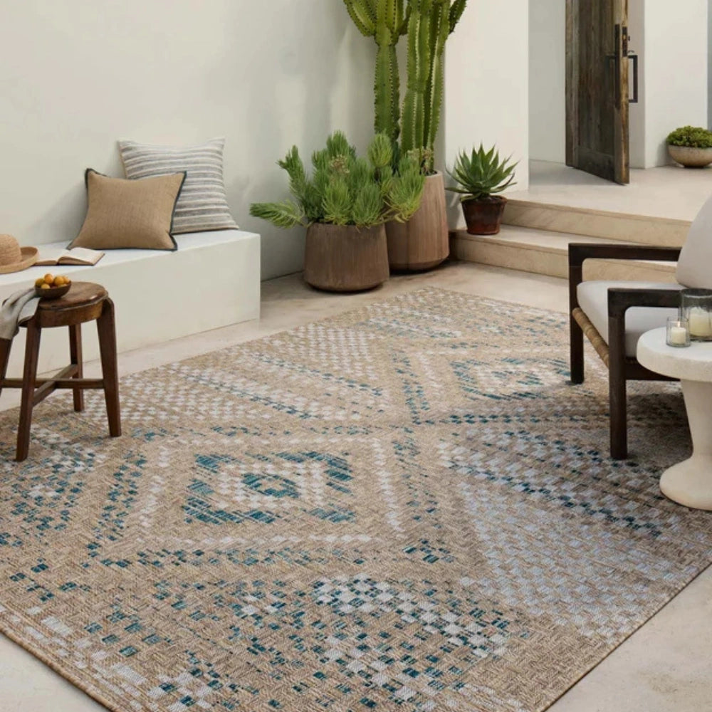 A large rectangular area outdoor rug with a bohemian-style geometric pattern in beige, blue, and white, styled in a bright, modern outdoor patio setting with plants, a bench, and a chair.
