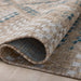 A close-up of a woven area outdoor rug with a rolled corner, showing a textured pattern in beige, cream, and blue on a neutral floor.