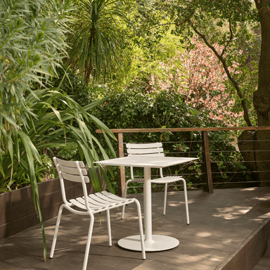 2 white armless stackable outdoor dining chairs and a square white dining table set on wooden deck patio surrounded by plants and trees