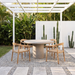 Outdoor dining set featuring a round concrete table and four light wood chairs with woven seats, arranged under a white pergola on a gravel patio with tropical plants and cacti in the background.