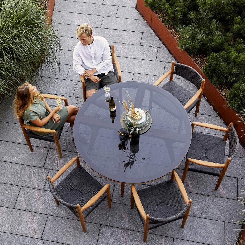 Boxhill's Aspect Teak Round Dining Table Clear Glass | Smokey Black lifestyle image with dining chairs and 2 people sitting down