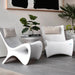 Two modern white sculptural in-pool lounge chairs with curved shell-like backs and bases, styled in a minimalist interior with gray tile flooring, a side table, books, and potted plants.