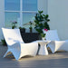 Two modern white molded outdoor in-pool lounge chairs with a small round white side table on a wooden deck, with potted plants and a window in the background.
