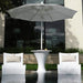 A modern outdoor patio scene with a large white patio umbrella centered over a white in-pool pedestal side table, flanked by two matching white in-pool lounge chairs with beige pillows, set beside a reflective pool in front of a building with dark glass windows.