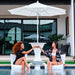 Two women sitting in white in-pool lounge chairs under a large white patio umbrella, toasting at a white in-pool pedestal table in a swimming pool. Modern glass-front building and outdoor furniture are visible in the background.