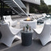 Modern outdoor patio furniture set with four white sculptural in-pool lounge chairs arranged around a round white fire pit table and a small dark gray side table, on a poolside deck with a staircase and pool in the background.