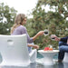 A woman sits in a white molded outdoor in-pool chair beside a small in-pool round table, holding a glass mug near another person's hand in a poolside setting with trees in the background.