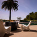 Modern outdoor patio scene with a round black fire pit table in the center, surrounded by sculptural white in-pool chairs, with a swimming pool, palm tree, and blue sky in the background.