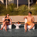 A man and a woman relax in white outdoor in-pool lounge chairs by a swimming pool, wearing swimwear and sunglasses, with a small white side table between them holding drinks. Tall green ornamental plants and a modern building are in the background.