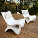 Two modern white sculptural in-pool lounge chairs on a wooden deck with trees in the background. Each in-poo white chair has a curved shell-like form and a striped throw pillow.