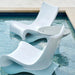 Two modern white curved outdoor in-pool lounge chairs beside a swimming pool, with a small matching in-pool side table between them.