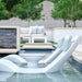 Modern white outdoor in-pool lounge chaise with matching round in-pool side tables and light blue patterned pillows, arranged in a swimming pool with a patio and lounge seating area in the background.