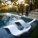 Two white wavy in-pool lounge chaise with a small in-pool round ice bin side table in the center, placed in a swimming pool outdoors near steps and trees at sunset.