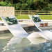 Two white outdoor in-pool lounge chaise with green tropical leaf print pillows are positioned in a swimming pool next to a small round in-pool side table holding two copper mugs. The background shows a green lawn, wooden fence, and stone wall.