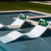 Two modern white curved outdoor in-pool lounge chaise with green tropical leaf print pillows, positioned next to a swimming pool with a small round in-pool resin side table between them.