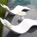 Two white wavy modern outdoor in-pool lounge chaise with light beige pillows and a matching small round in-pool side table, positioned beside a swimming pool on a dark deck.