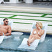A man and a woman relax in white in-pool lounge chaise in a swimming pool at a resort, with a small white in-pool side table between them.
