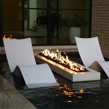 Modern outdoor fire pit with a long rectangular in-pool resin body and active flames, placed in a dark water feature beside two matching curved lounge chairs in a patio setting.