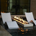 Modern outdoor fire pit with a long rectangular in-pool resin body and active flames, placed in a dark water feature beside two matching curved lounge chairs in a patio setting.