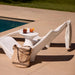 A white modern in-pool lounge chaise beside a swimming pool on a beige patio, with a small round in-pool side table, two drinks, a woven straw tote bag, and a striped towel draped over the backrest.