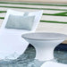 A modern outdoor poolside scene featuring a white lounge chair with a gray patterned pillow and a matching round pedestal side table in shallow water.