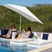 A large white patio umbrella shades two women relaxing on white and navy blue poolside lounge chairs by a swimming pool, with a lush green forest background.
