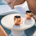 Two hammered copper Moscow mule mugs with gold-toned handles and stirrers, placed on a round in-pool white side table near a pool, with a person's legs and feet visible in the background.