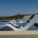 A row of modern white curved outdoor in-pool lounge chairs arranged around a pool, with a small round in-pool side table and potted pink flowers in the center. The background shows a scenic landscape with hills and a clear blue sky.