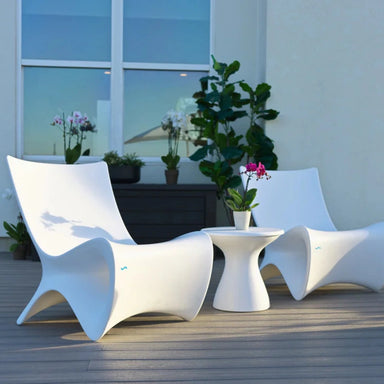 Two modern white  in-pool lounge chairs with a small white in-pool side table on a wooden deck, set against a light wall with potted plants and a window.