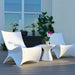 Two modern white molded in-pool lounge chairs with a small white in-pool side table on a wooden deck, set against a light wall with potted plants and a window in the background.