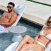 Two people relax on white lounge in-pool chairs by a swimming pool, with a small round in-pool side table between them. The setting is outdoors on a pool deck with a green strip of grass visible.