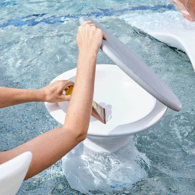 A white in-pool side table by the pool, with a person's arms reaching in and holding the lid open. The background shows blue swimming pool water.