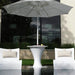 Outdoor patio furniture set with a white pedestal in-pool  side table under a large white umbrella, flanked by two white lounge chairs with beige pillows, set beside a reflective pool in front of a modern building.