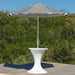 A black and white striped patio umbrella with a white pedestal table, set on a wooden deck with a lush green forest background.