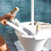 A white outdoor patio table with a built-in sink and lid, shown by a poolside. A person's hands are visible holding the lid open, and a beverage can and magazine are placed on the table.