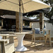 Outdoor patio furniture set including a white pedestal table, beige Adirondack-style chairs with blue pillows, and a large beige patio umbrella on a stone patio.