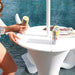 A white round outdoor in-pool  table by a pool with a person holding a beverage can beside it, another can on the table, and a folded chair nearby.