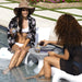 Two women relax by a swimming pool at a outdoor cafe setting. One woman is wearing a white bikini, a black and white floral open-front cardigan, and a brown wide-brim hat, holding a glass.