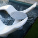 A modern white outdoor poolside chair with a circular seat and in-pool ice bin side table, positioned in dark pool water next to green grass.