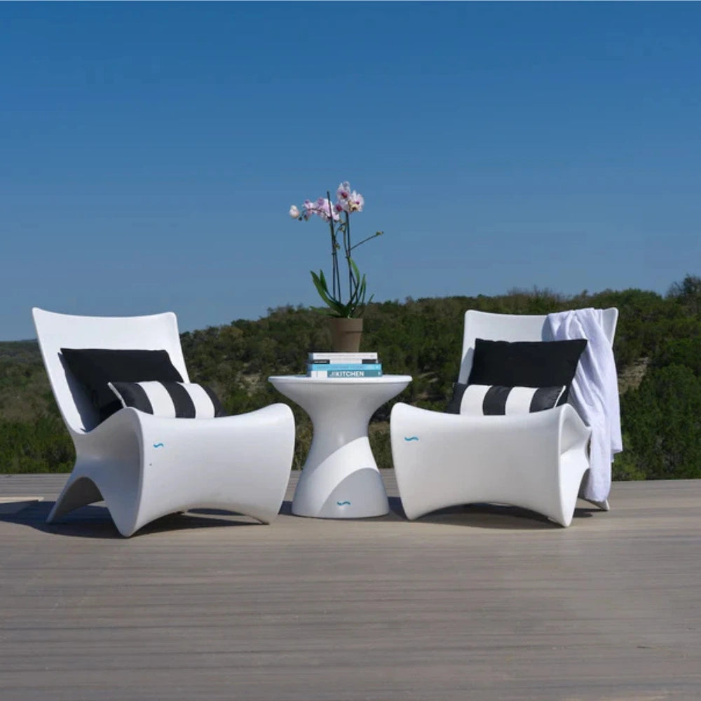 Two modern white lounge chairs with black and white striped pillows, a matching white side table, and a potted orchid on a wooden deck with a green landscape and blue sky in the background.