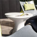 A white outdoor pedestal side table with a circular top and a built-in ice bin, shown next to modern gray lounge chairs with green patterned cushions.