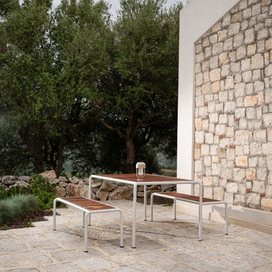 Patio scene with white steel framed rectangular dining table with wooden top and two matching benches against a stone wall surrounded by trees
